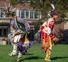 Two students dressed in Indigenous outfits perform a dance at a ceremony.