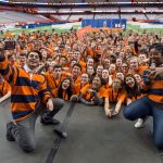 Syracuse University orientation leaders pose on stage with cameras to take a photo of students during orientation.