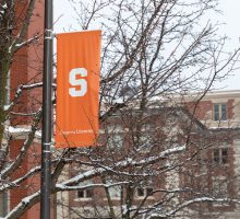A white block S on an orange banner hangs on a lamp post in front of Simms Hall