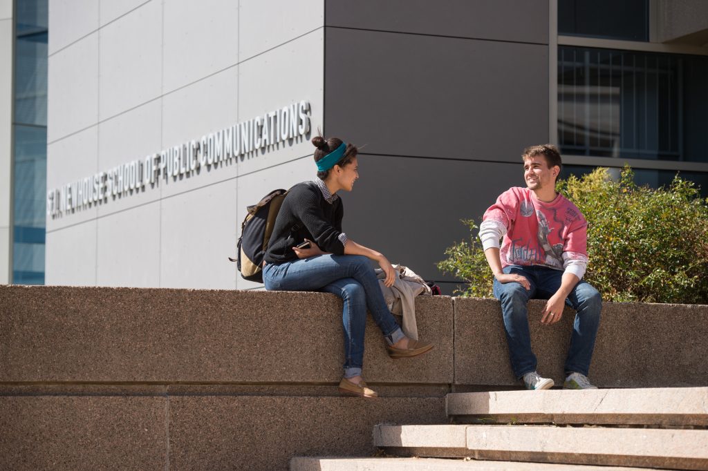 Two students sitting near the front steps of Syracuse University Newhouse School of Public Communications talking and connecting.