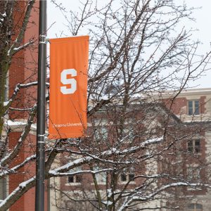 A white block S on an orange banner hangs on a lamp post in front of Simms Hall