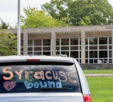 Van in front of residence hall. Back of van window has 'Syracuse Bound' in writing.