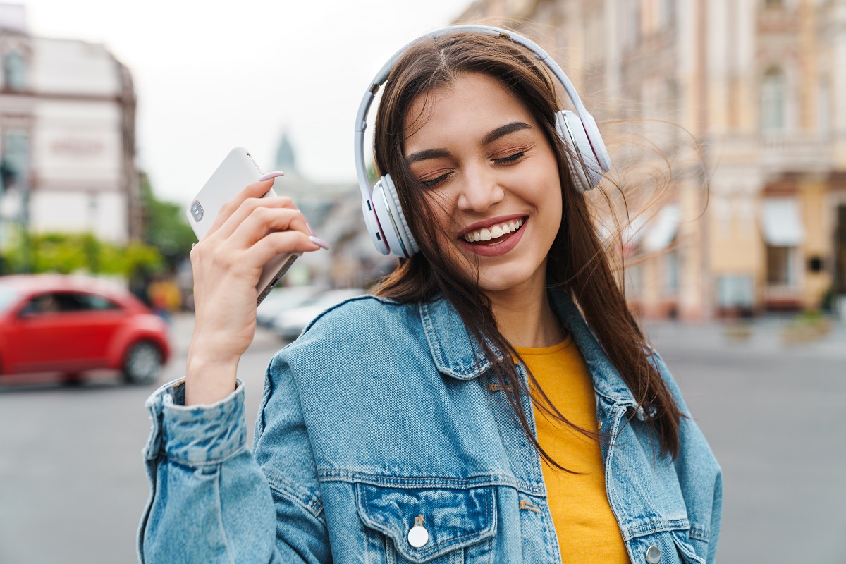 A woman happily listens to music on headphones on a street.