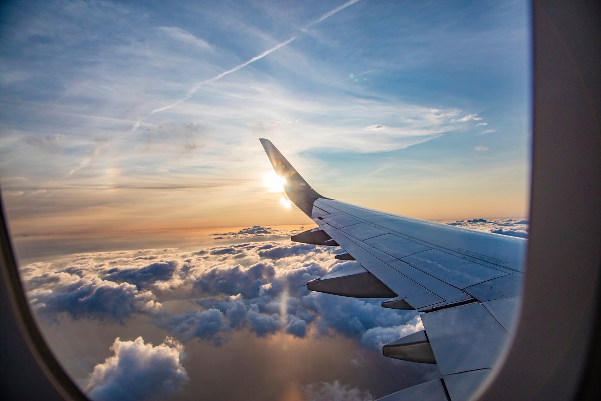 A photo of an airplane wing taken through an airplane window as the plane flies over some clouds and the sun sets.