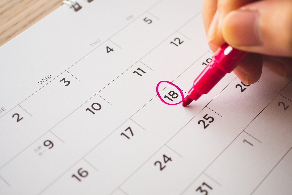 A hand holds a pink marker and circles the date of the 18th on a paper calendar.