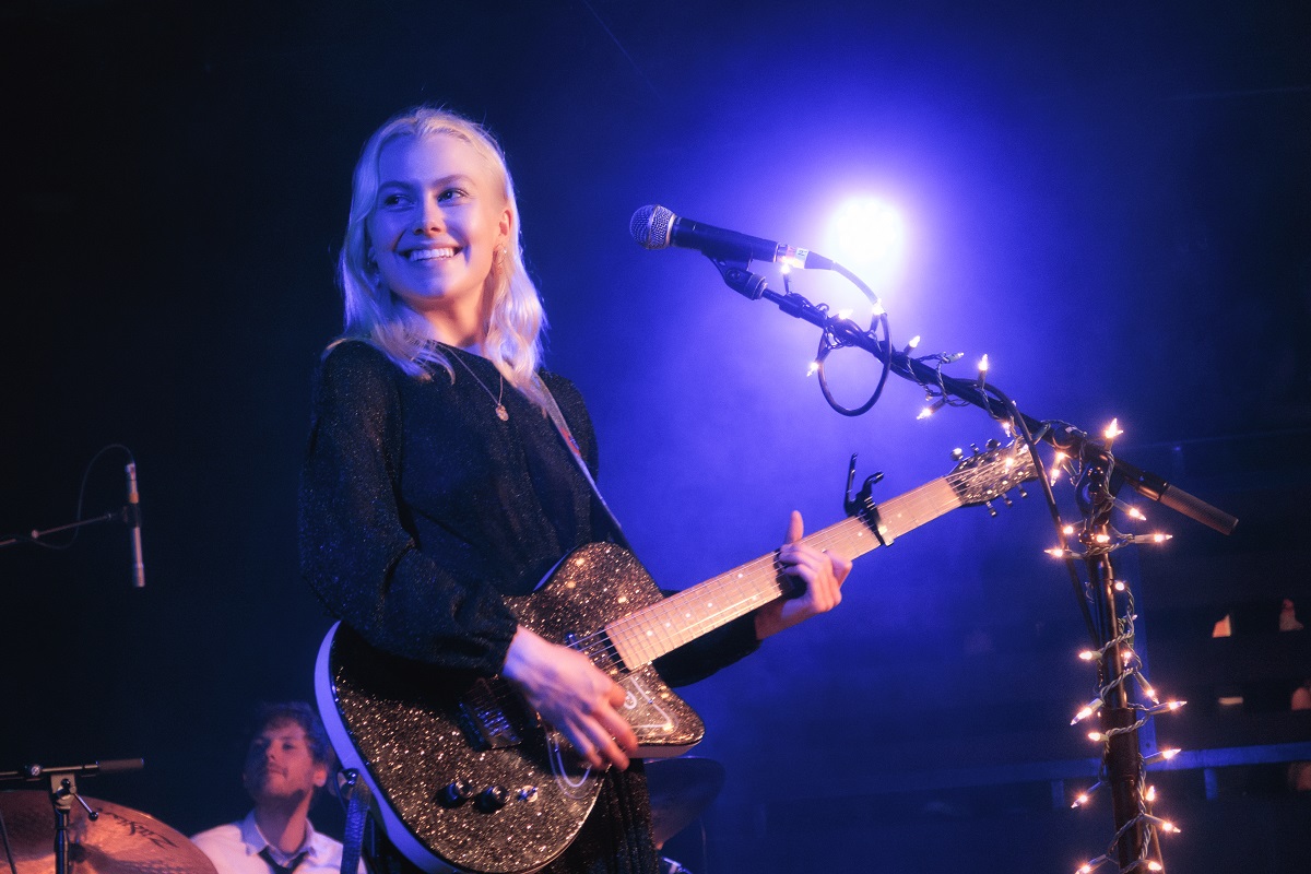 Phoebe Bridgers holds a guitar and smiles on stage