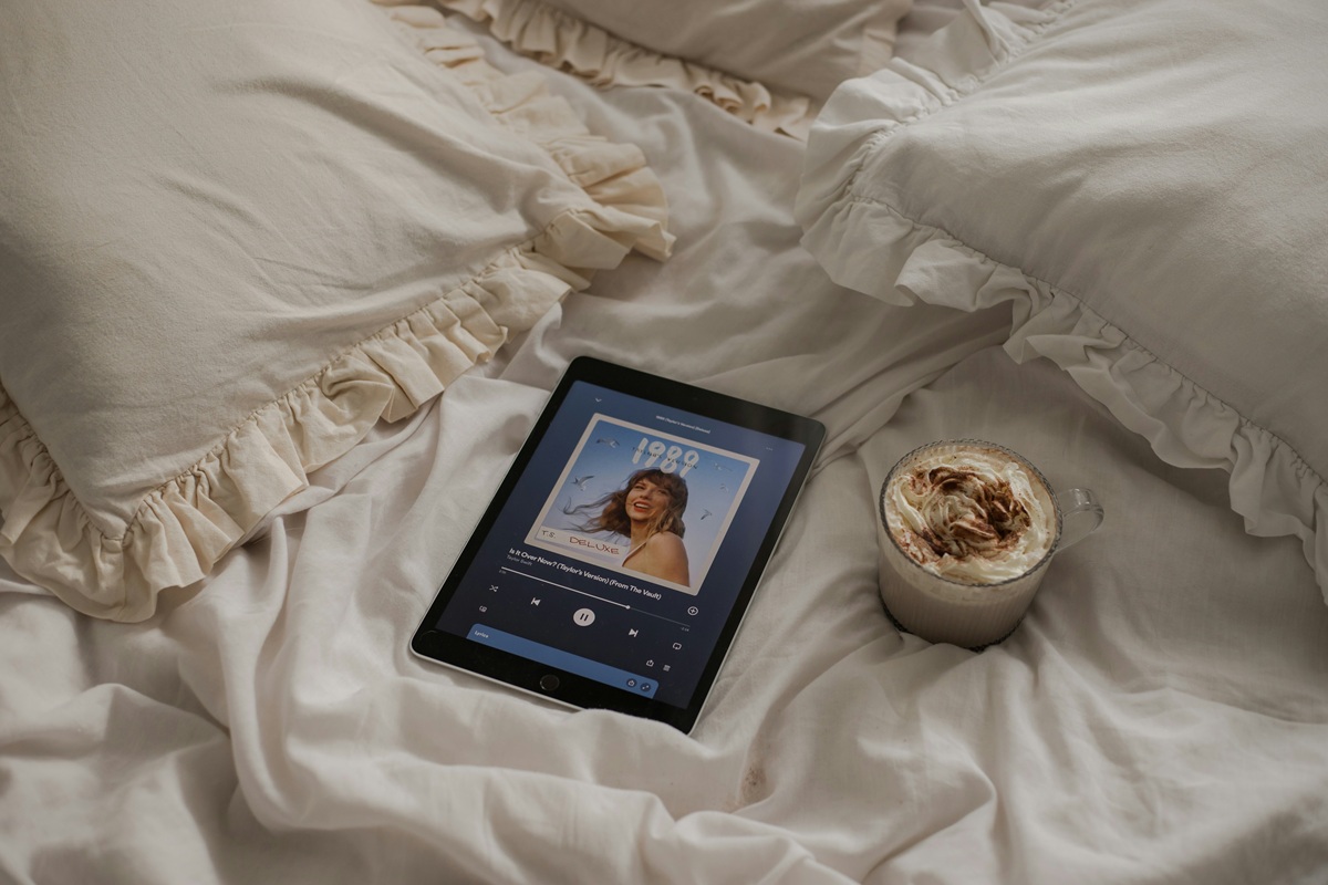 An iPad and a mug of hot chocolate with whipped cream sits on a bed. A song from "1989 (Taylor's Version" is visibly playing on the iPad screen.