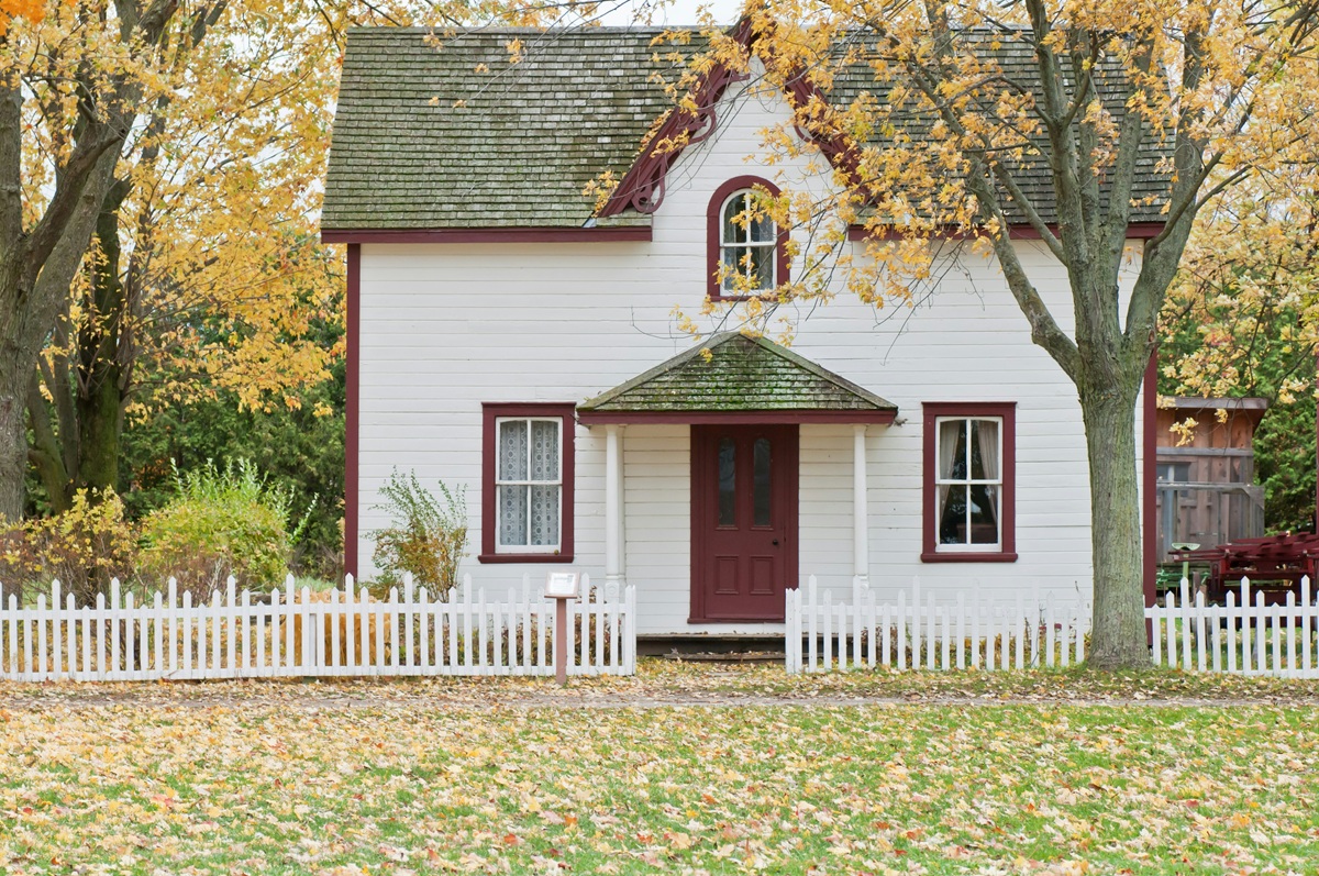 A white house with red trim surrounded by trees with yellow leaves and a white picket fence.
