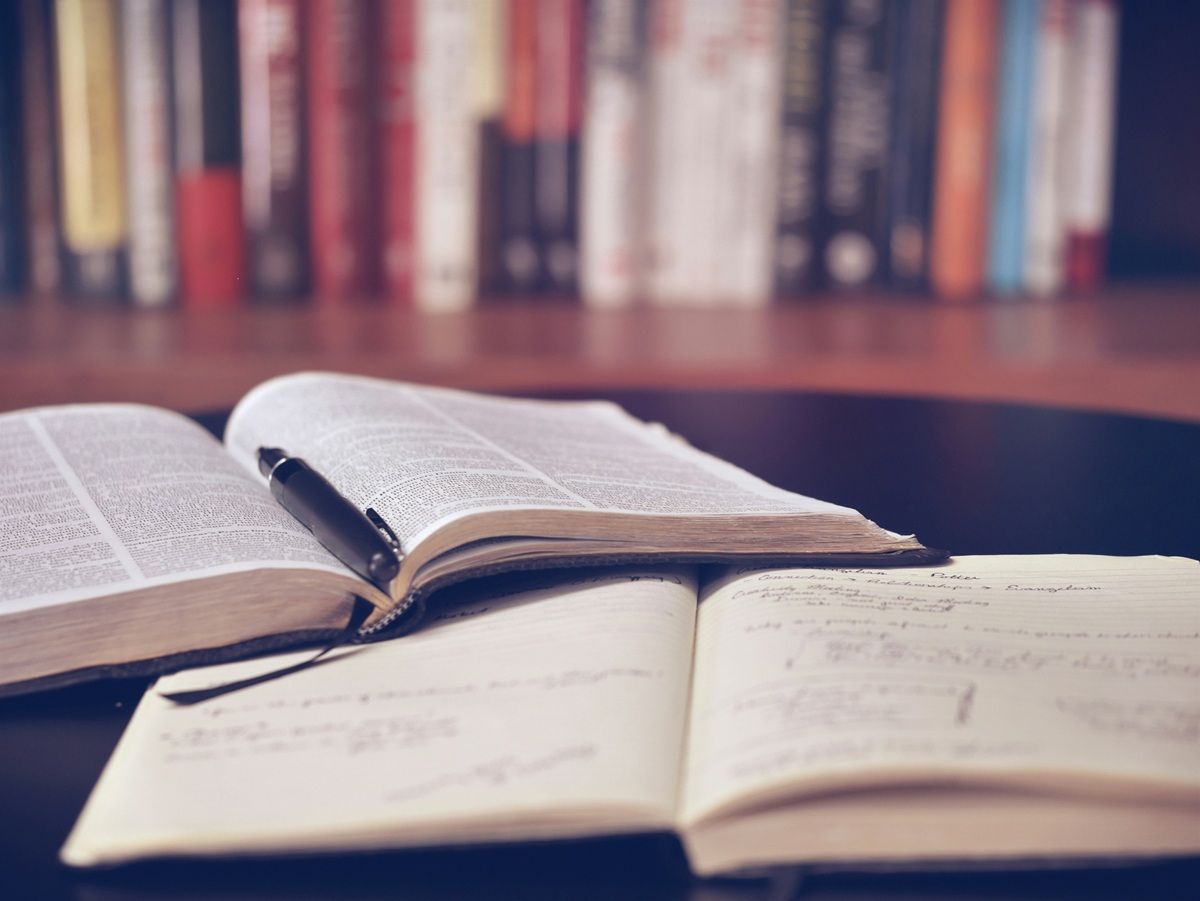 A book and a notebook lying open on a desk in front of a bookshelf.