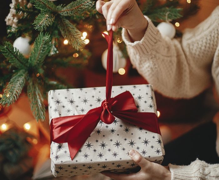A woman pulls a ribbon on a wrapped gift by a Christmas tree.