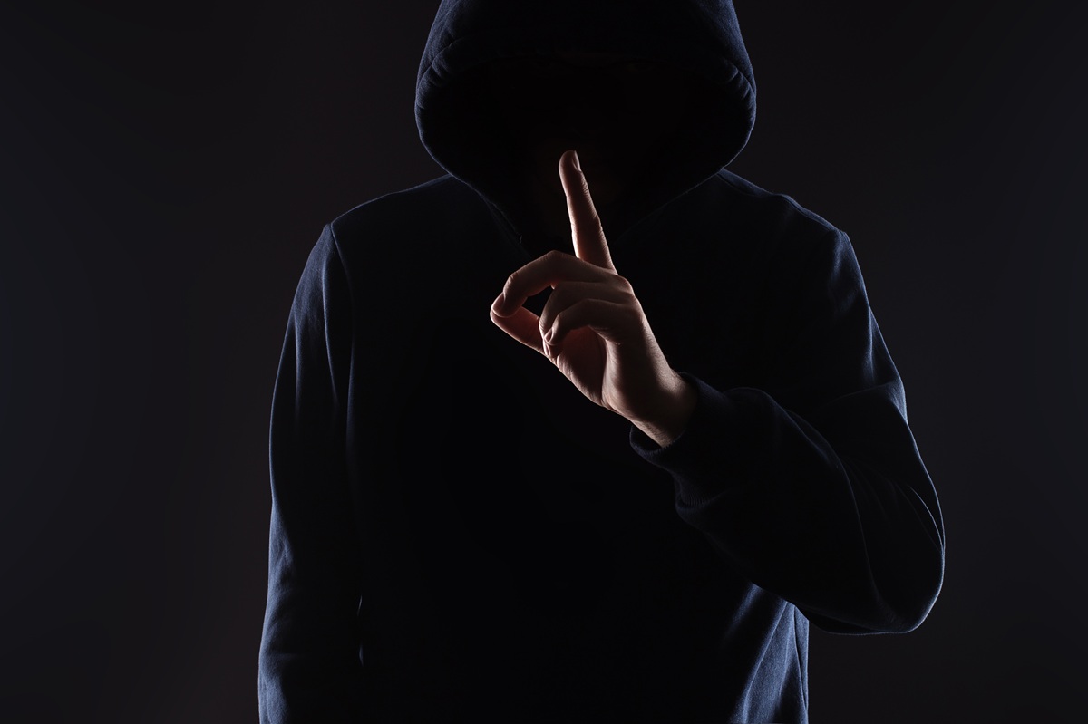 A person wearing a dark hoodie holding their index finger up to their lips.
