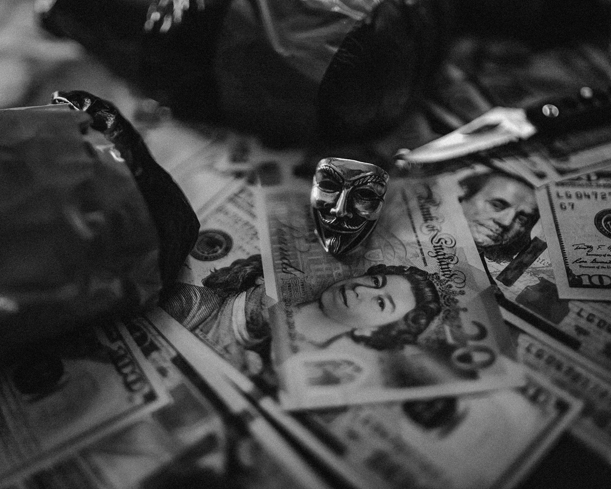 A pile of money from various countries is scattered across a table, along with a small knife, a silver Guy Fawkes mask figurine and various other indiscernible things.