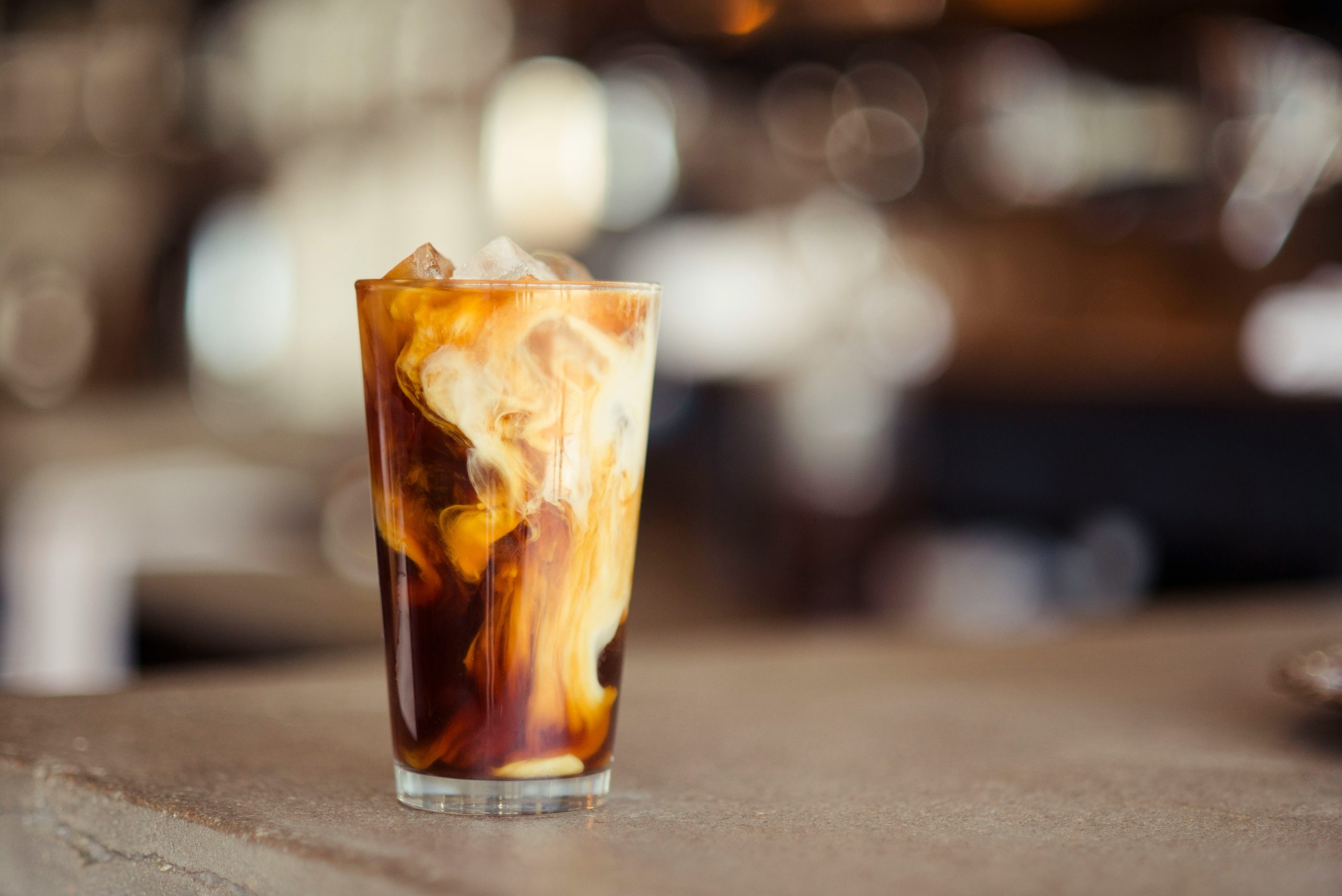 A glass of iced coffee with cream swirling through it on a café table.