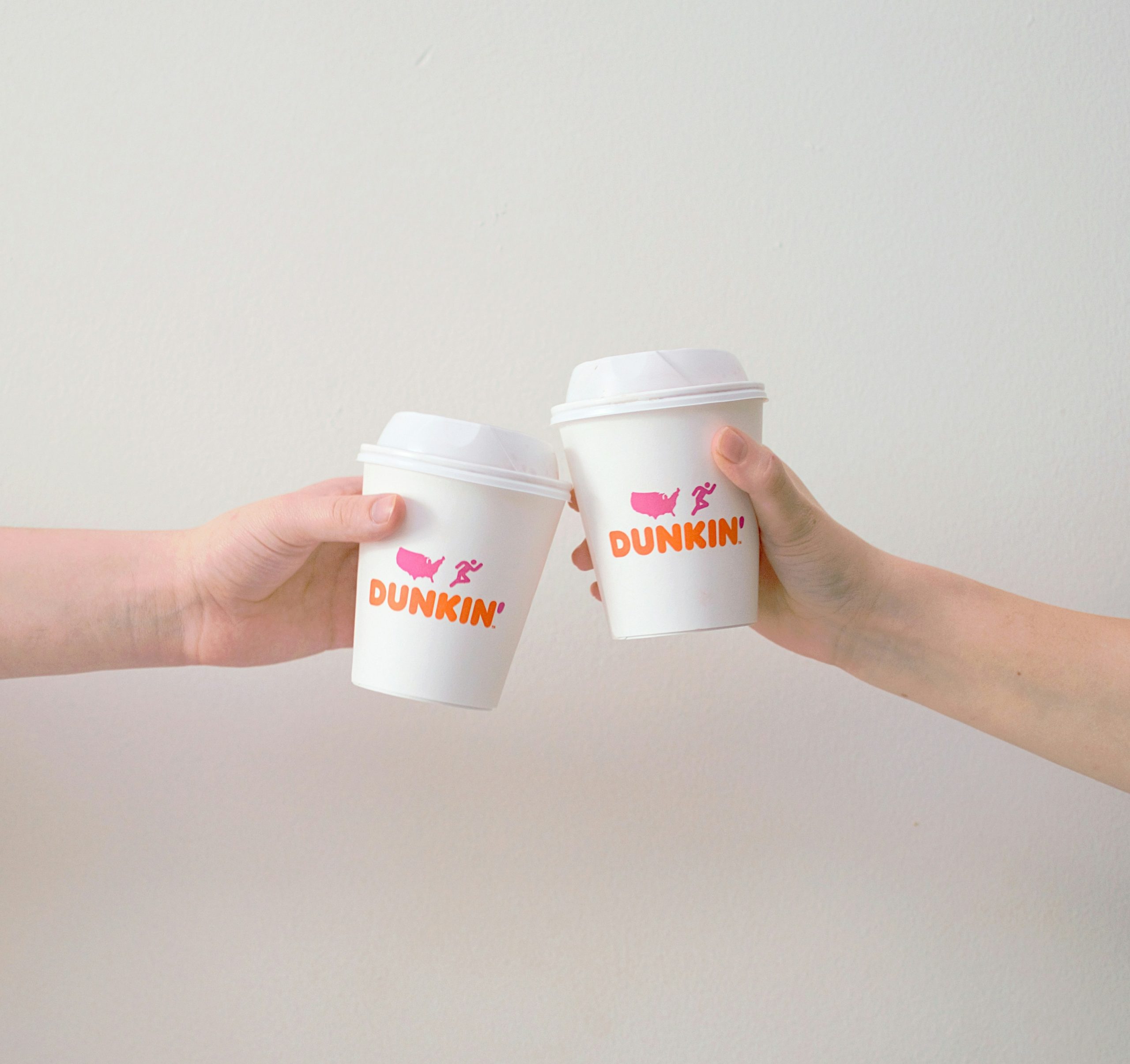 Two people hold up Dunkin’ coffee cups in a toast against a plain white background.