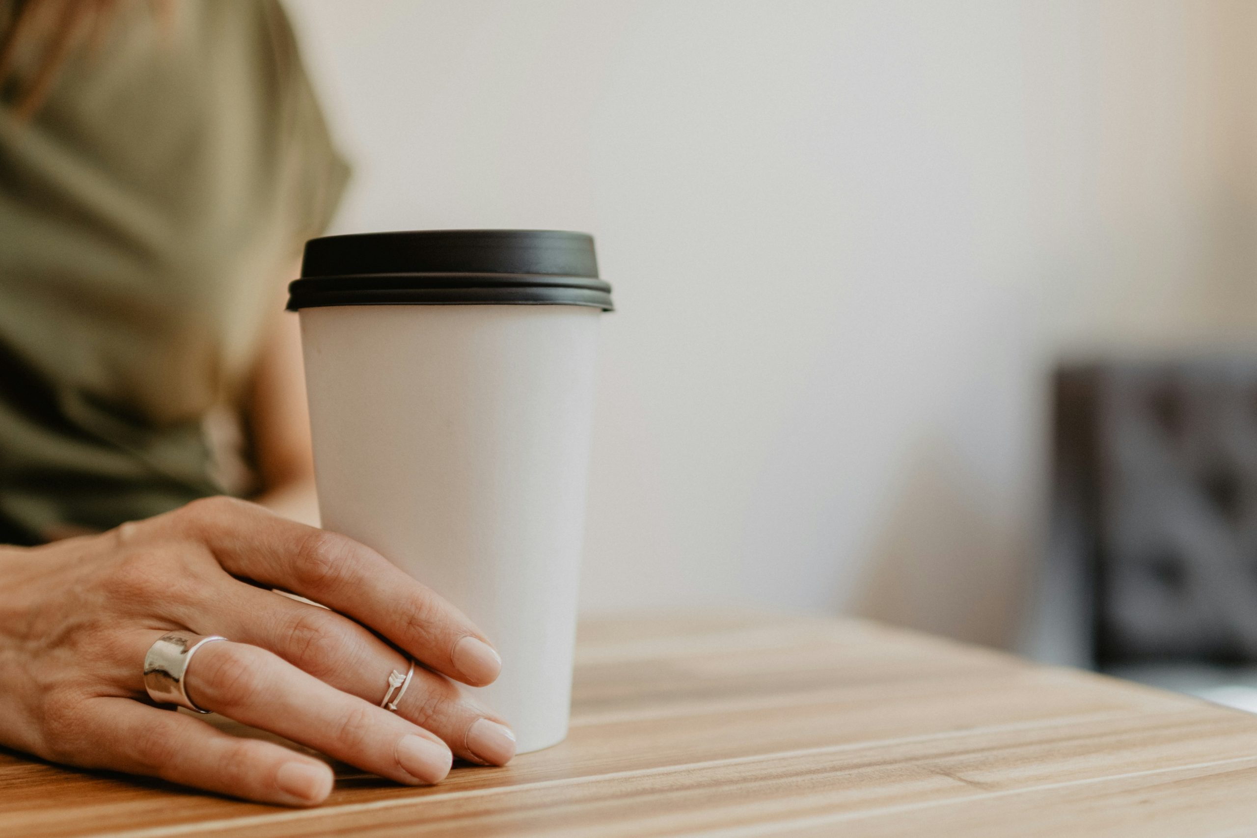 Close-up of a person’s hand resting beside a white takeaway coffee cup on a wooden table, with a softly blurred background.