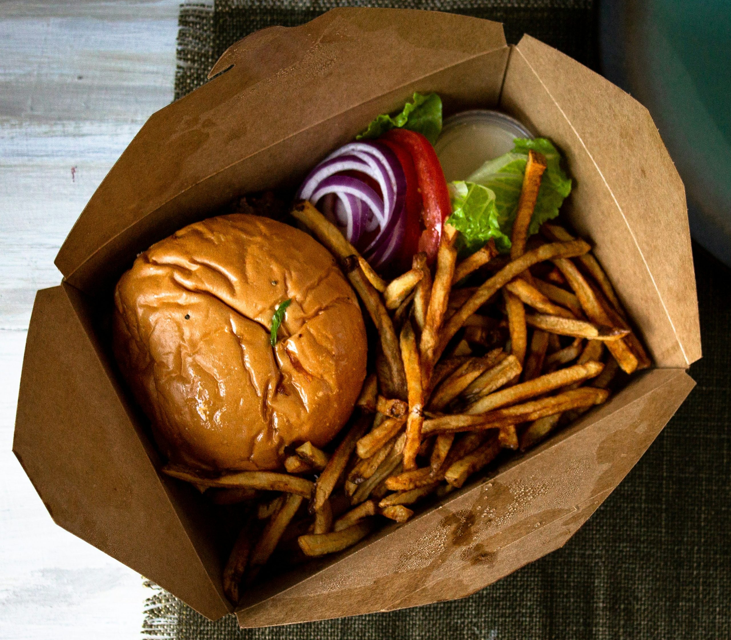 A burger topped with lettuce, tomato and red onion sits beside a serving of crispy fries in an open takeout box.