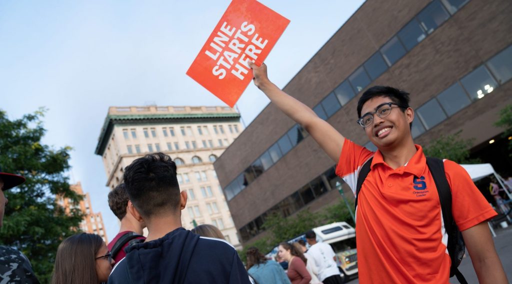 Syracuse University Orientation Leader smiling and holding up a sign that says line starts here.