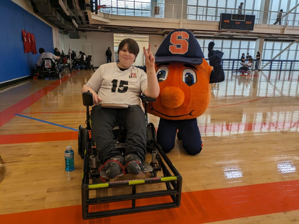 A student participating in OrangeAbility poses with Otto the Orange on the Barnes Center basketball courts